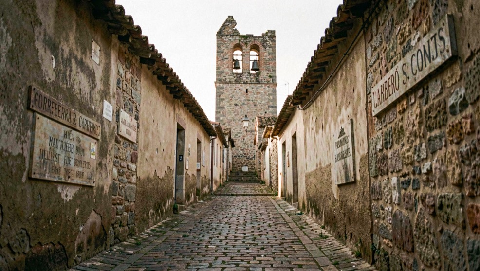 Vinyeta de la peça: Carreró empedrat que puja cap a un campanar truncat, rètols antics a les parets i llum baixa entre les teules.